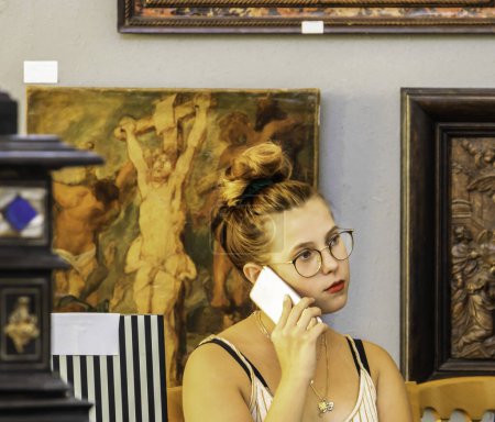 Foto de Cannes, Francia, 4 de agosto de 2019Bonita joven que llama con un teléfono inteligente mientras trabaja sentada en una sala de subastas durante una subasta de arte llamando a un cliente para una venta - Imagen libre de derechos