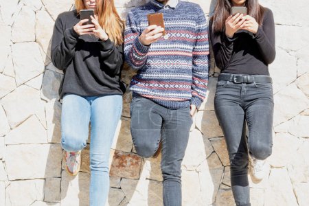 Young people using their smartphone lined up standing against a wall with their faces cut off unrecognizable person