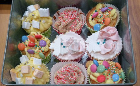 Téléchargez les photos : 9 cupcakes différents avec décoration : glaçage, bonbons à la guimauve, crème, glaçage royal, pâte à sucre sculptée en forme d'arc et de rose, perles de sucre dans une boîte à étain vintage - en image libre de droit