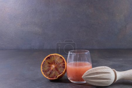 Foto de Jugo recién exprimido de naranja sangre en un vaso, con media naranja sangre y un exprimidor de mano de madera, sobre un fondo liso con espacio negativo para el texto - Imagen libre de derechos