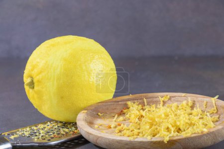 Zitrone auf einer Stahlreibe, sowie ein Holzteller mit der Zitronenschale, die von der Schale der Zitrusfrucht entfernt wurde.