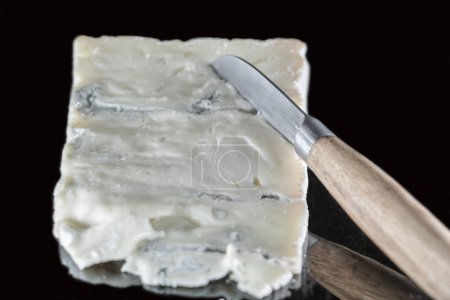Italian Gorgonzola cheese on a black background with mirror effect and a cheese knife with a wooden handle resting on it, different layers of blue and cream visible