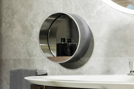 Photo for Detail on the round mirror of a modern steel bathroom above the sink basin with a bottle of perfume for men - Royalty Free Image