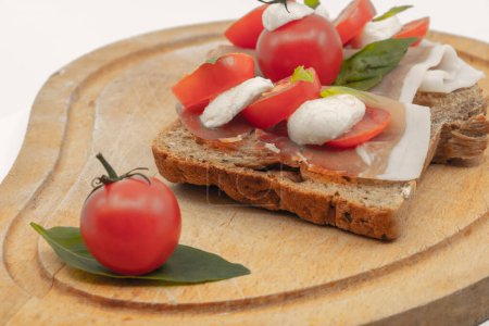 Téléchargez les photos : Sandwichs italiens sur une tranche de pain multi-grains avec une tranche de tomates cerises jambon rouge boules de mozzarella et basilic frais sur une assiette en bois - fond blanc isolé - en image libre de droit