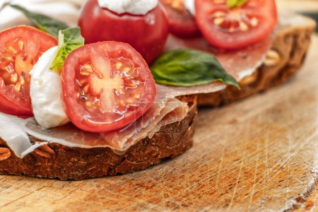 Foto de Sandwiches italianos en una rebanada de pan de grano múltiple con una rebanada de jamón rojo tomates cherry bolas de mozzarella y albahaca fresca en un plato de madera - fondo blanco aislado - Imagen libre de derechos