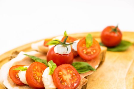Téléchargez les photos : Sandwichs italiens sur une tranche de pain multi-grains avec une tranche de tomates cerises jambon rouge boules de mozzarella et basilic frais sur une assiette en bois - fond blanc isolé - en image libre de droit