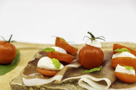 Foto de Sandwiches italianos en una rebanada de pan de grano múltiple con una rebanada de jamón rojo tomates cherry bolas de mozzarella y albahaca fresca en un plato de madera - fondo blanco aislado - Imagen libre de derechos