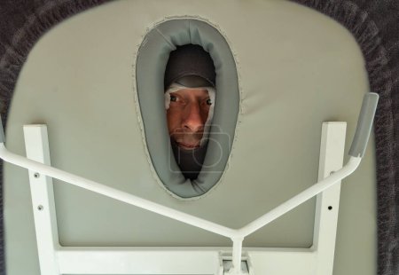 Patient at the physiotherapist during a massage - face of man seen from under the massage table by the hole of the head rest at the masseur at the hospital or rehabilitation center