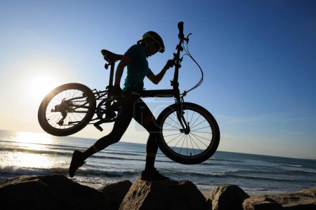 Téléchargez les photos : Femme portant un vélo pliant au lever du soleil bord de mer - en image libre de droit
