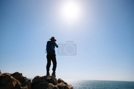 Téléchargez les photos : Femme photographe prenant des photos sur les rochers littoraux du lever du soleil - en image libre de droit