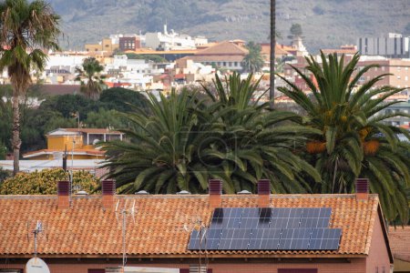 Foto de Paneles solares en el techo de una casa unifamiliar de ladrillos rojos y tejas rojas, con palmeras y paisaje de árboles en el fondo. Tenerife, Islas Canarias, España - Imagen libre de derechos