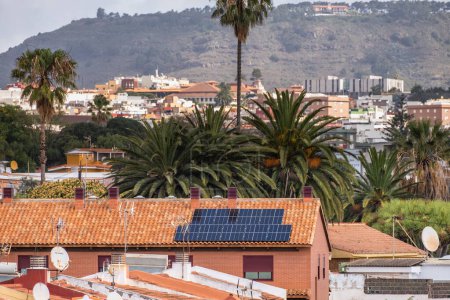 Foto de Paneles solares en el techo de una casa unifamiliar de ladrillos rojos y tejas rojas, con palmeras y paisaje de árboles en el fondo. Tenerife, Islas Canarias, España - Imagen libre de derechos