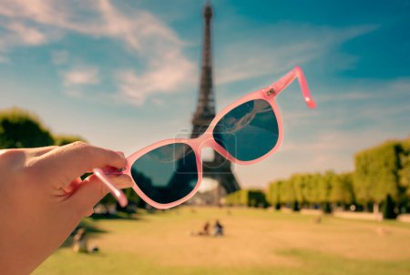 Photo for Paris - pink sunglasses in the hand of a tourist. Eiffel Tower and Field of Mars landscape as background. Travel poster - summer vacation (holiday) in Europe. - Royalty Free Image