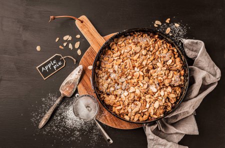 Photo for Homemade apple pie with flaked almonds crumble. Cake in a baking tin. Dessert captured from above (top view, flat lay) on a black background. Free copy (text) space. - Royalty Free Image