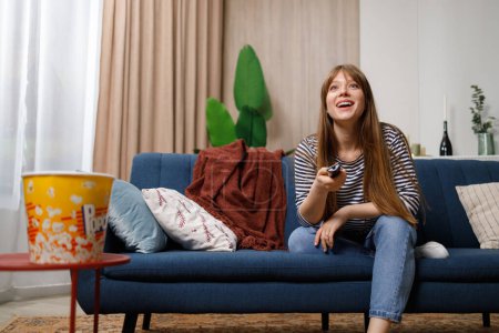Glücklich überraschte junge Frau mit Fernbedienung in der Hand fernsehen, während sie auf dem Sofa sitzt