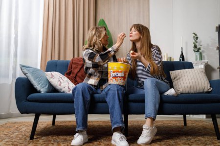 Foto de Madura madre y su hija adulta alimentándose mutuamente con palomitas de maíz sentado en el sofá en acogedor apartamento - Imagen libre de derechos