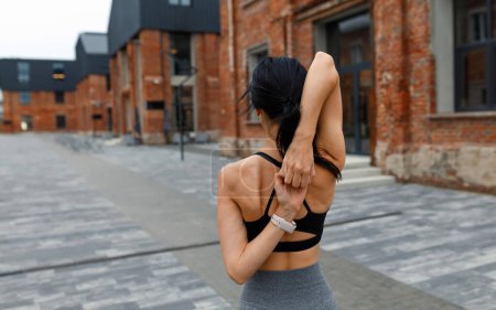Athletic woman exercising yoga pose and clasping hands behind her back on lock, rear view against brick building Poster 620436020