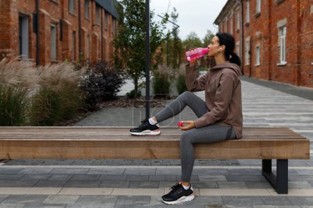 Photo for Sports woman in earphones drinking water after jogging on the bench - Royalty Free Image