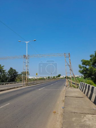 Téléchargez les photos : Stock photo of empty asphalt road surrounded by green plants and trees, LED street lamps or light in the middle of the road, blue sky on background. Picture captured under bright sunlight at Gulbarga. - en image libre de droit