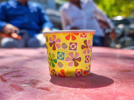 Téléchargez les photos : Stock photo of empty yellow color floral design printed paper cup kept on red color plastic table, blurred people in background. Picture capture under bright light at Gulbarga, karnataka, India. - en image libre de droit