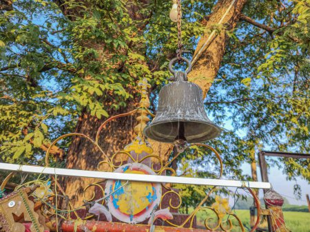 Photo for Stock photo of ancient copper or bronze bell hanging on a iron frame at Hindu temple, green tree on background. Picture captured under bright sunlight at Tuljapur road, Maharashtra, India. - Royalty Free Image