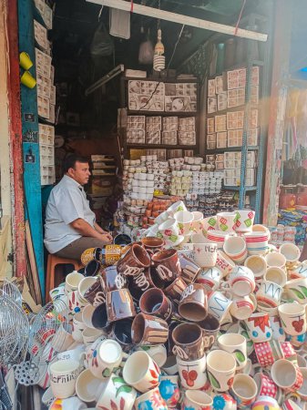 Téléchargez les photos : Gulbarga, Inde - 26 novembre 2022 ; Photo de stock de 40 à 50 vendeur indien âgé assis à son magasin et vendant coloré, tasse de thé et café en céramique concepteur et tasses dans la zone du marché. - en image libre de droit