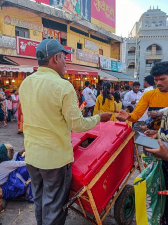 Photo for Tuljapur,India- October 28th 2022; Stock photo of Adult Indian man wearing yellow shirt, gray pant and cap, selling ice cream outside of the temple area. hotel, shop, crowd of devotees on background. - Royalty Free Image