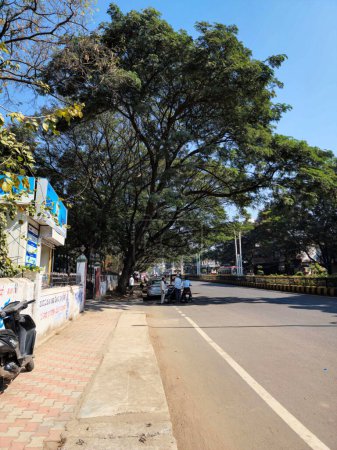 Téléchargez les photos : Kalburgi, Inde - 18 janvier 2023 ; photo d'un sentier ou d'un sentier où peu de gens se parlent, mendiant assis sur le sol. Il y a une voiture blanche garée, quelques grands arbres verts entourant - en image libre de droit