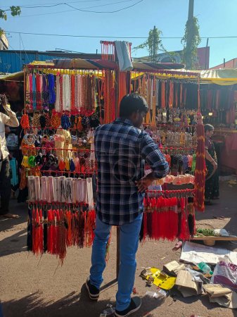 Téléchargez les photos : Tuljapur, Inde- 28 octobre 2022 ; photo de stock de jeune garçon indien debout au milieu de la route sur le marché vendant bande religieuse colorée poignet, collier aux clients, sous la lumière du soleil - en image libre de droit