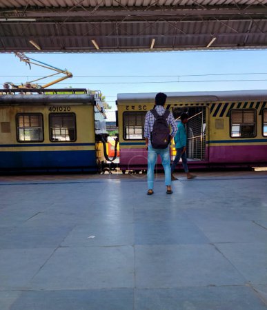 Foto de Hyderabad, India- 12 de octubre de 2022; El joven, vestido casual y luciendo una mochila negra, se paró en la plataforma del ferrocarril, mirando al tren local en el fondo, esperando su llegada.. - Imagen libre de derechos