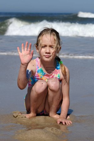The child sits on the background of the Atlantic Ocean and waves his hand at the camera. A seven-year-old girl sits on the beach and smiles.