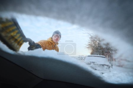 Photo for Man clearing snow from windshield his snowy car after snowfall. Parking on city street in winter - Royalty Free Image