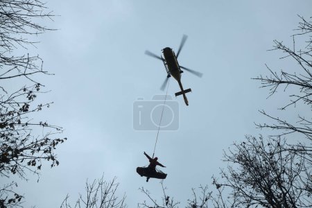 Photo for Emergency service paramedic with patient suspended by rope under helicopter. Rescue from difficult to access terrain - Royalty Free Image