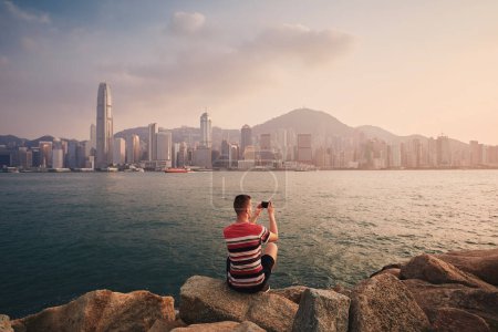 Téléchargez les photos : Rear view of man photographing Hong Kong urban skyline at sunset. Tourist using smart phone in modern city - en image libre de droit