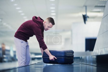 Foto de Traveling by airplane. Passenger pick up his blue suitcase in baggage claim in airport terminal - Imagen libre de derechos