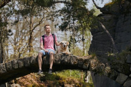 Photo for Man with dog on hike in the middle of rocks. Pet owner is sitting on stone bridge together with loyal labrador retriever. - Royalty Free Image
