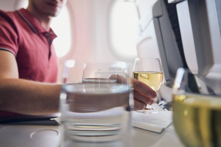 Téléchargez les photos : Man enjoying drink during flight. Passenger holding glass of white wine against airplane window - en image libre de droit