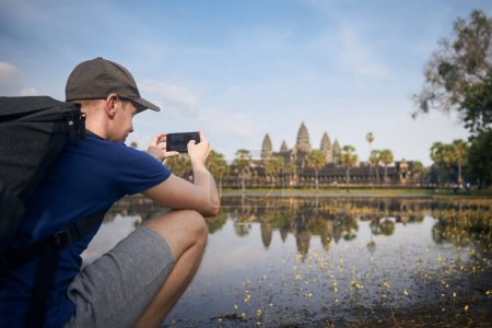 Photo for Man with backpack taking pictures of water reflection of temple in lake. Tourist in Angkor Wat near Siem Reap in Cambodia - Royalty Free Image