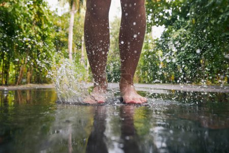 Téléchargez les photos : Homme sautant avec les pieds nus dans la flaque pendant la journée pluvieuse. Gros plan des jambes et éclaboussures d'eau - en image libre de droit
