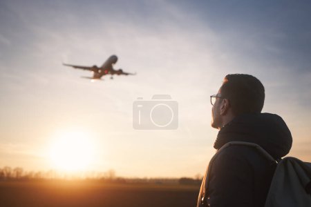Photo for Man with backpack looking up to airplane landing at airport during beautiful sunset. - Royalty Free Image