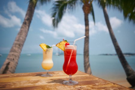 Foto de Dos cócteles bajo palmeras en la hermosa playa. Singapur Sling y Pina Colada bebidas sobre mesa de madera. - Imagen libre de derechos