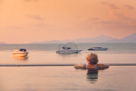 Téléchargez les photos : Femme âgée active dans la piscine. Vue arrière de la femme appréciant se détendre et regarder la mer au coucher du soleil - en image libre de droit
