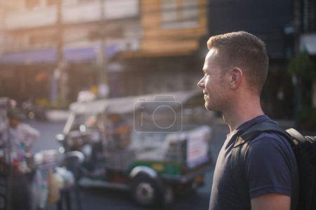 Photo for Portrait of tourist with backpack walking at street. Side view of man against tuk tuk on road. Bangkok, Thailan - Royalty Free Image