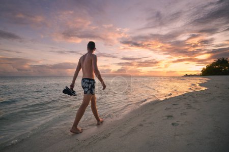 Téléchargez les photos : Vue arrière de l'homme en maillot de bain tenant des tongs à la main, marchant pieds nus seul sur une plage de sable idyllique et regardant le coucher de soleil coloré sur l'océan - en image libre de droit