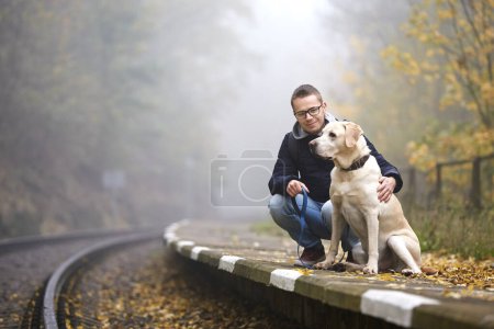 Foto de El hombre con el perro en la correa están esperando en la plataforma de la estación de tren. Propietario de mascotas y su labrador recuperador juntos durante el viaje en tren - Imagen libre de derechos