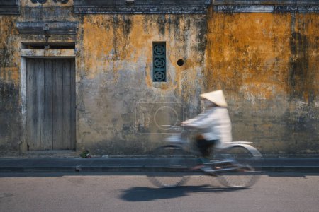 Vietnamese woman with traditional hat riding bicycle in old town. City life in Hoi An, Vietnam