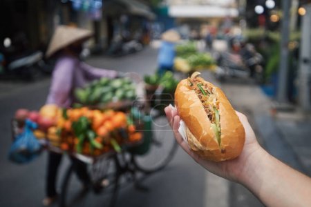 Foto de Comida callejera en Hanoi. Mano sosteniendo Banh Mi sándwich. Primer plano de la baguette tradicional vietnamita llena de paté, carne y verduras - Imagen libre de derechos