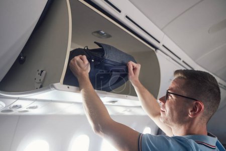 Photo for Man travel by airplane. Passenger putting hand baggage in lockers above seats of plane - Royalty Free Image