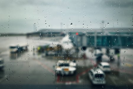 Téléchargez les photos : Météo sombre à l'aéroport. Concentration sélective sur les gouttes sur la fenêtre du terminal contre l'avion sous la pluie - en image libre de droit
