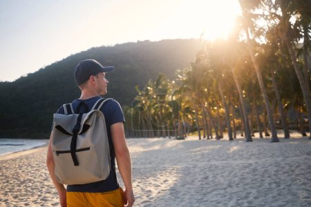 Téléchargez les photos : Vue arrière du touriste avec sac à dos marchant sur une belle plage de sable avec des palmiers et regardant le coucher du soleil. - en image libre de droit